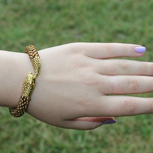 May include: A gold-colored bracelet in the shape of a snake, with a detailed scale pattern. The bracelet is wrapped around a wrist. The nails are painted a light purple color.