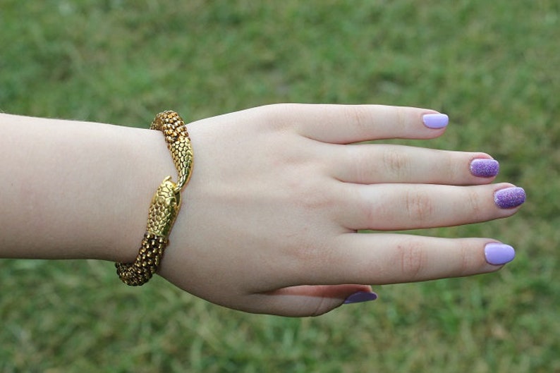May include: A gold-colored bracelet in the shape of a snake, with a detailed scale pattern. The bracelet is wrapped around a wrist. The nails are painted a light purple color.