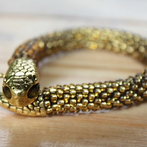May include: A gold-colored snake bracelet with a detailed snake head. The bracelet is made of small, round beads, creating a textured, scale-like appearance. The snake's head has intricate detailing.