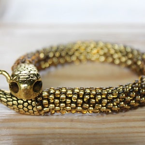 May include: Gold-toned snake bracelet. The bracelet features a snake head with detailed scales and a curved body. The body of the bracelet is made of small, gold-colored beads. The bracelet is on a wooden surface.