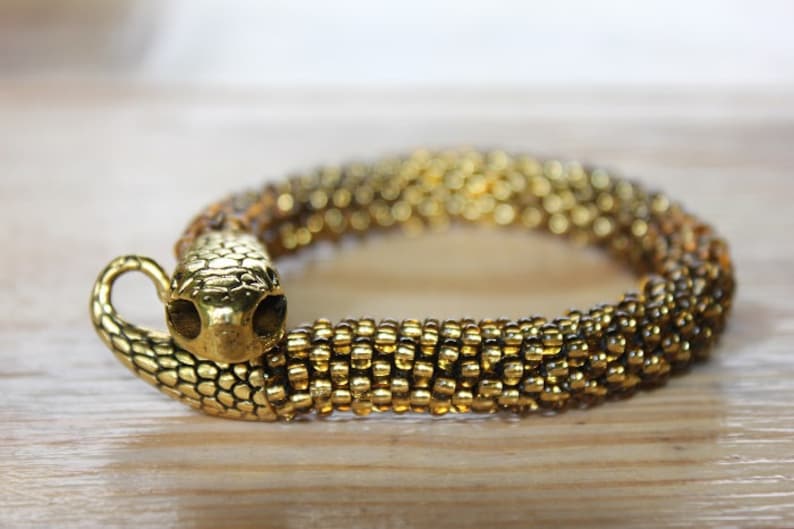 May include: Gold-toned snake bracelet. The bracelet features a snake head with detailed scales and a curved body. The body of the bracelet is made of small, gold-colored beads. The bracelet is on a wooden surface.