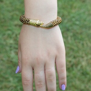May include: A gold-toned snake bracelet with textured scales. The bracelet is wrapped around a wrist, with the snake's head and tail meeting at the clasp. The background is blurred green grass.