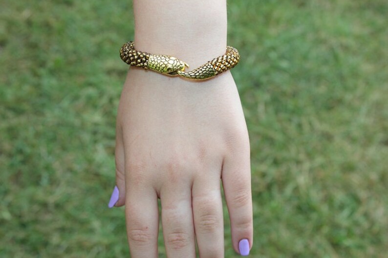 May include: A gold-toned snake bracelet with textured scales. The bracelet is wrapped around a wrist, with the snake's head and tail meeting at the clasp. The background is blurred green grass.