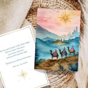 Puede incluir: Tarjeta de Navidad con una pintura de acuarela de tres figuras montadas en camellos hacia una estrella. La tarjeta incluye texto con borde dorado e ilustración de estrella. El fondo muestra montañas y una puesta de sol.
