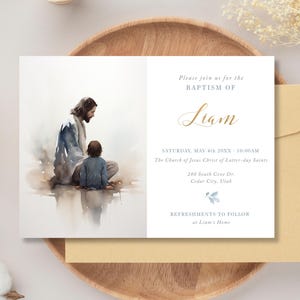 May include: Baptism invitation featuring a watercolor illustration of Jesus with a child. The invitation includes the name "Liam" in gold script, event details, and the church address. The invitation is on a white background with a tan envelope.