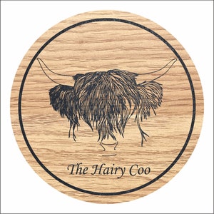 May include: A round wooden coaster featuring a black and white illustration of a Highland cow's head. The text "The Hairy Coo" is printed below the illustration.