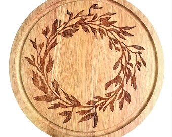 May include: A circular wooden serving board featuring an engraved leaf wreath design. The board is light brown with a darker brown engraved pattern. The edge has a carved groove. Suitable for serving cheese, appetizers, or as a decorative item.