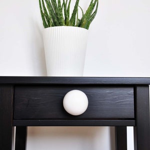 Peut inclure: Une table d'appoint en bois noir avec un bouton en céramique blanche sur le tiroir. Un pot en céramique blanche avec une plante d'aloe vera verte est posé sur la table.