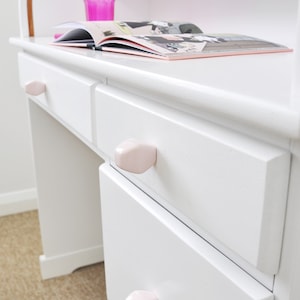 Puede incluir: Escritorio blanco con una parte superior curva y tres cajones. Los cajones tienen pomos de cerámica rosa. Un libro está abierto en el escritorio y un vaso rosa está encima.