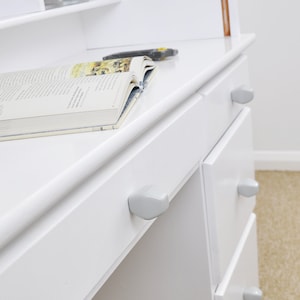 May include: White desk with two drawers and a shelf above. The drawers have gray knobs. A book is lying on the desk.