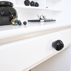 Könnte beinhalten: Weißer Schreibtisch mit einem schwarzen Schubladenknopf und einer schwarzen Keramikvase mit einer Halskette darauf. Der Schreibtisch hat eine weiße Tischplatte und weiße Beine. Auf dem Schreibtisch liegen ein Buch und eine Brille.