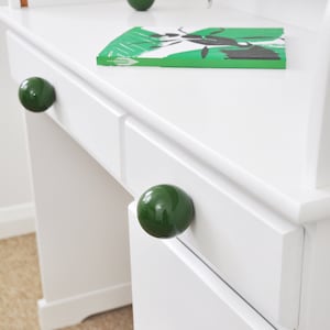 May include: White desk with three drawers and green round knobs. The top drawer is open and a book is on top of the desk.