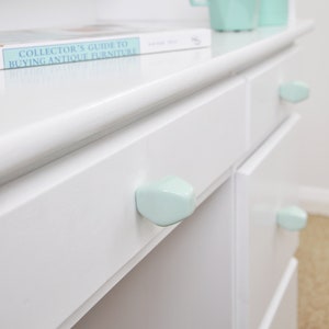 Puede incluir: Cómoda blanca con tiradores de cajones de cerámica azul claro. La cómoda tiene un libro titulado "Guía del coleccionista para comprar muebles antiguos" en la parte superior.