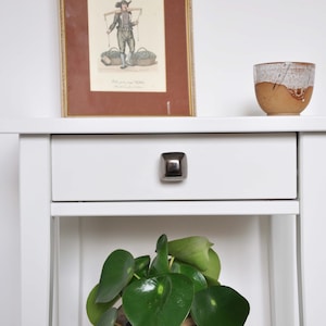 Puede incluir: Mesa auxiliar blanca con un solo cajón y un estante inferior. El cajón tiene una perilla cuadrada plateada. Una pequeña planta en maceta se encuentra en el estante inferior. Un cuadro enmarcado y una taza de cerámica están en la parte superior de la mesa.