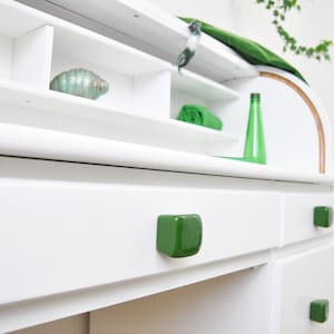 Puede incluir: Escritorio blanco con tres cajones y tiradores de cajones de cerámica verde. El escritorio tiene una parte superior curva y estantes abiertos con detalles verdes.