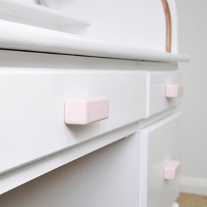 Puede incluir: Cómoda blanca con tres cajones y tiradores rectangulares de color rosa.