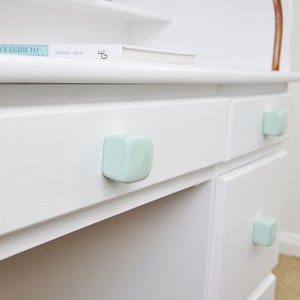 May include: White desk with three drawers and light blue ceramic knobs. The top drawer has a book titled "Collector's Guide To" on top.