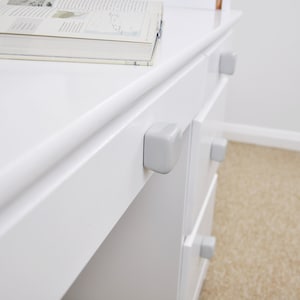 Könnte beinhalten: Weiße Kommode mit vier Schubladen und grauen Kunststoff-Schubladenknöpfen. Die Kommode ist aus Holz und hat eine glatte, weiße Oberfläche.