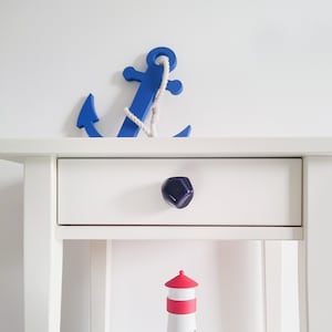 May include: White nightstand with a drawer and a purple knob. A blue wooden anchor is on top of the nightstand. A white lighthouse with a red base is on the bottom shelf.