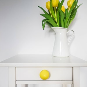 May include: A white nightstand with a single drawer and a yellow knob. A white pitcher with a bouquet of yellow tulips sits on top of the nightstand.