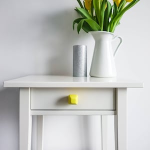 Puede incluir: Una mesa auxiliar blanca con un cajón y una perilla amarilla. Una vela plateada y una jarra blanca con tulipanes amarillos están encima de la mesa.