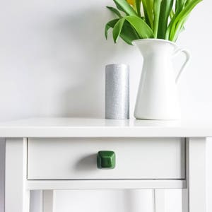 May include: A white nightstand with a single drawer and a green ceramic knob. A silver candle holder and a white pitcher with yellow tulips are on top of the nightstand.