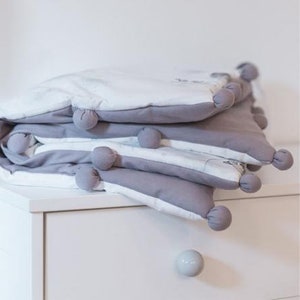 Puede incluir: Una manta de bebé gris y blanca doblada con pompones grises en los bordes. La manta está apilada sobre una cómoda blanca. La pared detrás de la manta tiene una calcomanía de luna y el texto "Let's fly to the moon".