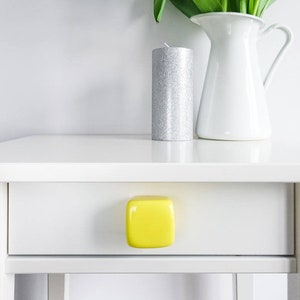 May include: A white bedside table with a yellow square drawer knob. A silver candle and a white jug with yellow tulips are on top of the table.