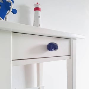 May include: White bedside table with a single drawer and a blue knob. The drawer is closed. A small white lighthouse figurine is on top of the bedside table. A white sign with blue lettering reads "BEACH" with a sun and waves graphic.