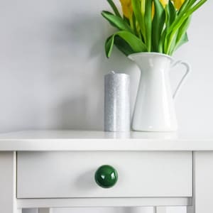 Może przedstawiać: Biała szafka nocna z jedną szufladą i zielonym ceramicznym gałką. Srebrna świeca i biały dzbanek z żółtymi tulipanami stoją na szafce nocnej.