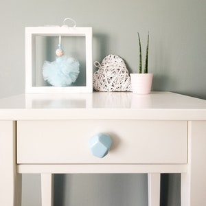 Puede incluir: Mesita de noche blanca con un solo cajón y una perilla geométrica azul claro. El cajón está cerrado. Encima de la mesita de noche hay un corazón de mimbre blanco, una pequeña planta suculenta en maceta y un marco de fotos blanco con una bailarina de tul azul dentro.