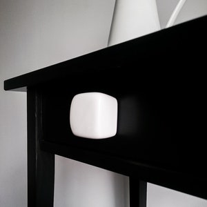 May include: A black wooden side table with a white ceramic drawer pull. The table has a simple design and is ideal for a small space.