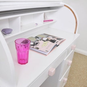 Puede incluir: Escritorio de madera blanco con un diseño de rollo superior. El escritorio tiene un vaso rosa en la superficie, un libro y tres cajones con pomos rosas. El escritorio tiene un estante sobre la superficie con un bolígrafo rosa y un pequeño objeto decorativo.