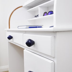 May include: White wooden desk with three drawers and a hutch. The drawers have blue ceramic knobs. The hutch has shelves and a curved top.
