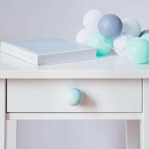 May include: A white bedside table with a drawer and a light blue knob. A white book is on top of the table, and a string of white and light green cotton ball lights is on the right side of the book.