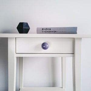 May include: White nightstand with a single drawer and a purple knob. A book with black text is on top of the nightstand, along with a dark gray geometric object.