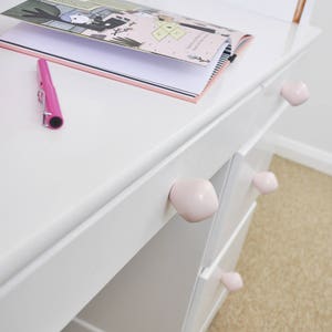 May include: White desk with three drawers and pink ceramic knobs. A pink pen and a closed book are on the desk.