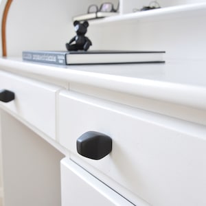 May include: White desk with two drawers and black drawer pulls. The desk has a top shelf with a book and a small black bear figurine.