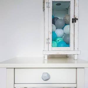 Puede incluir: Una mesita de noche blanca con un solo caj&oacute;n y un tirador de caj&oacute;n de piedra gris. Una linterna blanca con un frente de vidrio est&aacute; encima de la mesita de noche. La linterna est&aacute; llena de bolas de algod&oacute;n azules y grises.