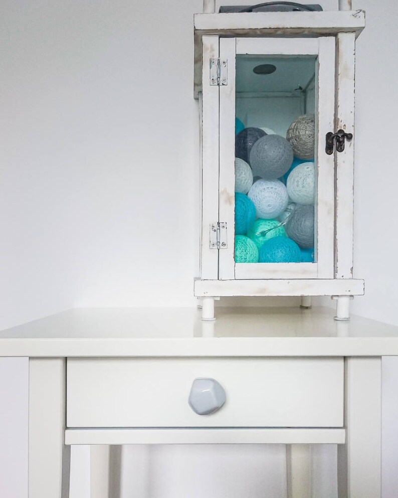 Puede incluir: Una mesita de noche blanca con un solo caj&oacute;n y un tirador de caj&oacute;n de piedra gris. Una linterna blanca con un frente de vidrio est&aacute; encima de la mesita de noche. La linterna est&aacute; llena de bolas de algod&oacute;n azules y grises.