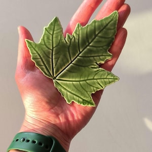 May include: A ceramic leaf in shades of green, with detailed veining, is held in a hand. The leaf's edges are slightly raised, giving it a three-dimensional appearance. The hand has a green watch strap.