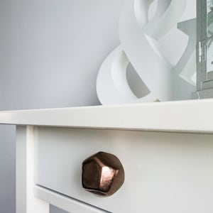 Puede incluir: Un cajón de cómoda blanco con una perilla geométrica de color cobre.