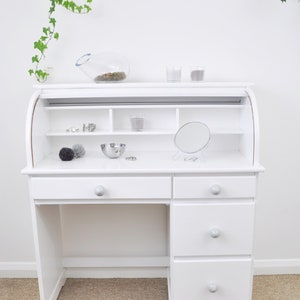 May include: White roll top desk with three drawers and a shelf above the drawers. The desk has a small round mirror and a bowl on the top surface.