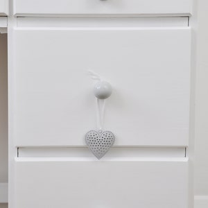 Pu&ograve; includere: Un com&ograve; in legno verniciato di bianco con tre cassetti. Ogni cassetto ha un pomello rotondo grigio. Un ornamento grigio a forma di cuore con un design traforato &egrave; appeso al cassetto centrale.