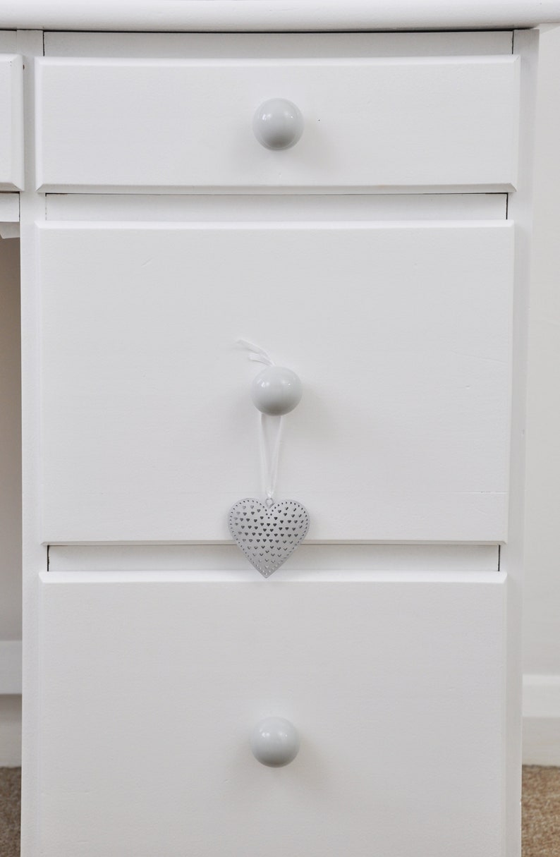 Pu&ograve; includere: Un com&ograve; in legno verniciato di bianco con tre cassetti. Ogni cassetto ha un pomello rotondo grigio. Un ornamento grigio a forma di cuore con un design traforato &egrave; appeso al cassetto centrale.
