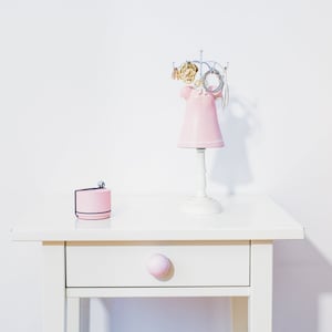 May include: A white table with a pink drawer and a pink ceramic jewellery stand with gold and silver jewellery. The stand is on the table with a pink box next to it.