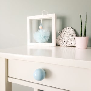 Könnte beinhalten: Eine weiße Kommode mit einem hellblauen Keramik-Knopf am Schubfach. Ein weißer Bilderrahmen mit einer blauen Tüllpuppe, die darin hängt, steht auf der Kommode. Eine weiße, herzförmige gewebte Dekoration und eine Topfpflanze stehen ebenfalls auf der Kommode.