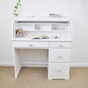 Pu&ograve; includere: Scrivania bianca a rullo con cassetti e ripiani. La scrivania ha un piano curvo e una superficie di scrittura estraibile. Un piccolo peluche, un libro aperto e alcuni piccoli oggetti sono sulla scrivania. La stanza ha pareti bianche e un tappeto beige.