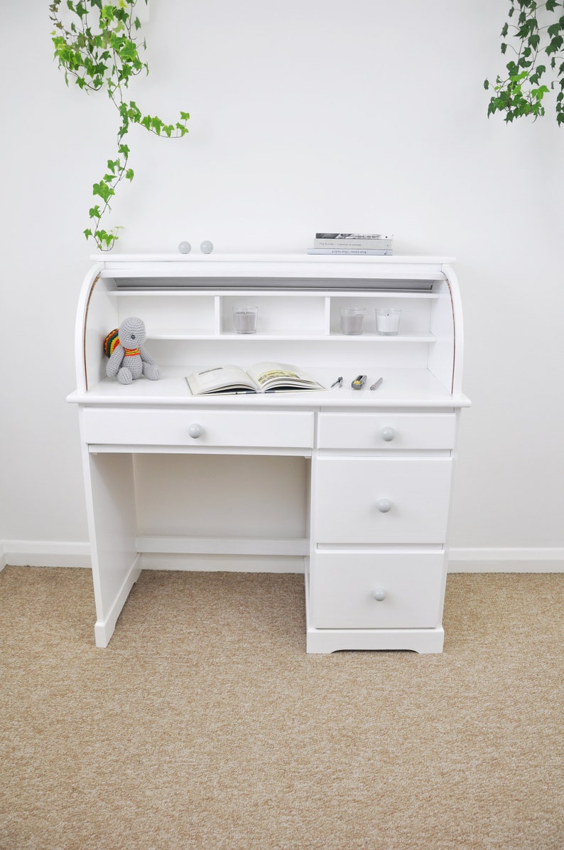 Pu&ograve; includere: Scrivania bianca a rullo con cassetti e ripiani. La scrivania ha un piano curvo e una superficie di scrittura estraibile. Un piccolo peluche, un libro aperto e alcuni piccoli oggetti sono sulla scrivania. La stanza ha pareti bianche e un tappeto beige.