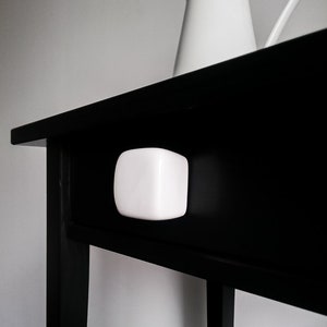 Puede incluir: Una mesa negra con un tirador de cajón blanco y cuadrado. Una jarra blanca con asa está sobre la mesa. La mesa tiene un diseño sencillo y minimalista.